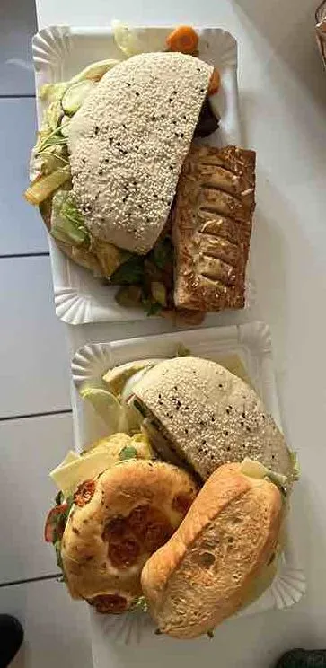 Zwei Pappteller mit belegten Brötchen und Gebäck auf einem weißen Tisch. Auf jedem Teller liegen verschiedene Sandwiches mit Salat sowie ein zusätzliches Brötchen oder Gebäckstück. Im Hintergrund sind Teile des Raums und Schuhe am Boden zu sehen.
