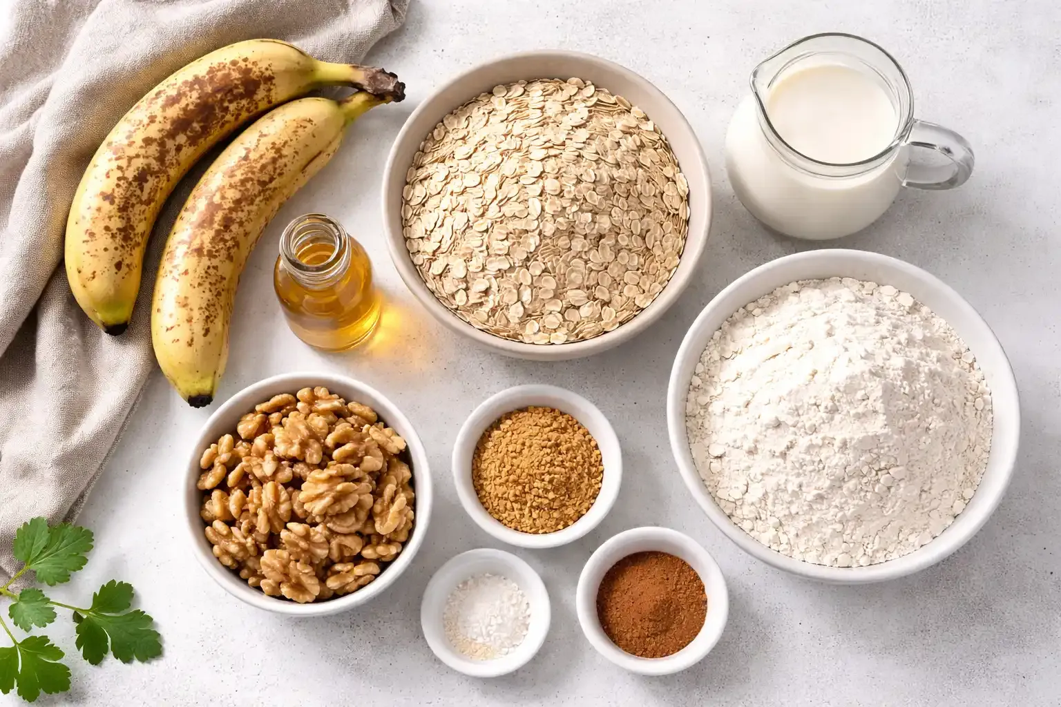 Draufsicht auf überreife Bananen, Haferflocken, Mehl, Pflanzenmilch und Walnüsse, die zusammen mit weiteren Backzutaten übersichtlich auf einer hellen Oberfläche angeordnet sind.